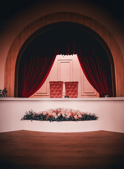 A stage set with a classic theatre design featuring a semi-circular wooden arch and deep red velvet curtains draped to frame two tufted red upholstered chairs centered behind a white panel backdrop. I