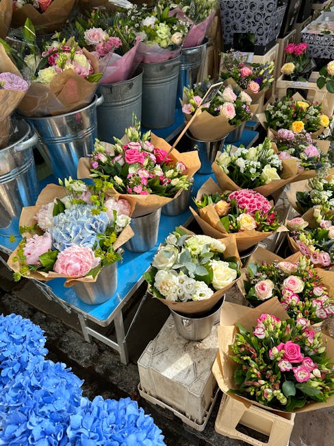 A vibrant display of freshly arranged floral bouquets featuring pastel pink roses, white chrysanthemums, light blue hydrangeas, and assorted greenery, arranged in small brown kraft paper wraps and pla